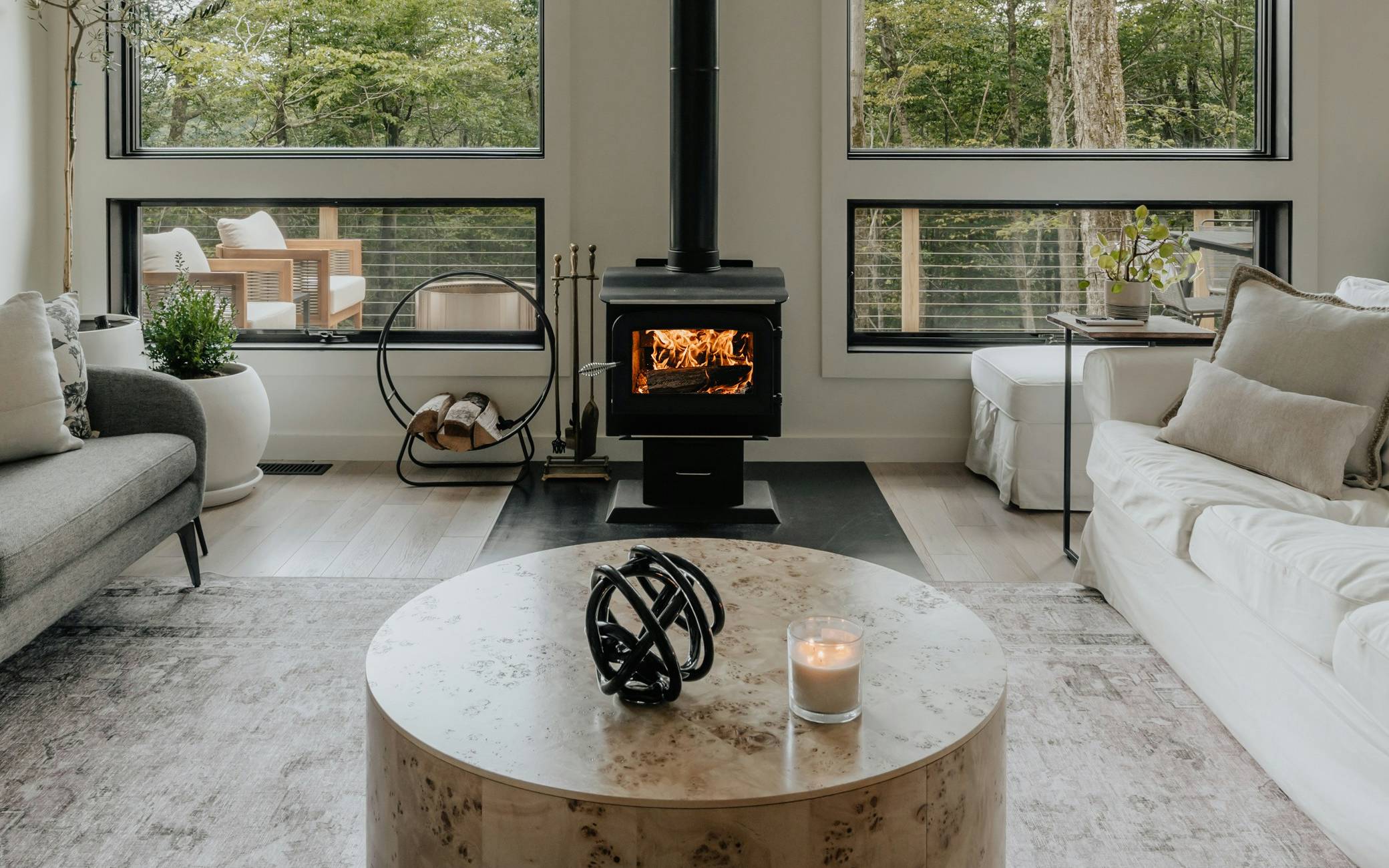 Living room with a wood stove between tall windows onto trees, flanked by grey and white sofas